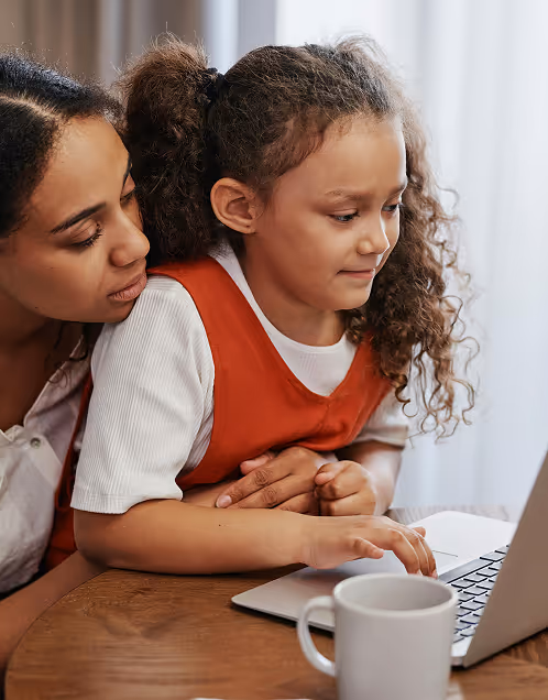 Woman helping a young girl with curly hair use a laptop at a wooden table.