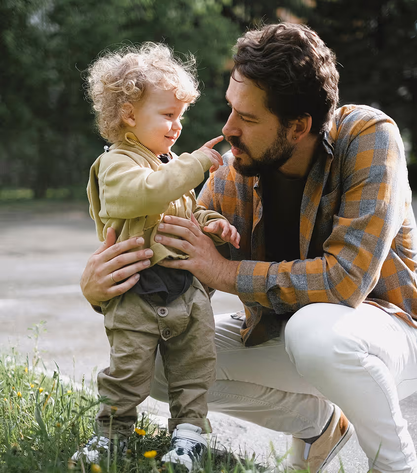 Man crouching and smiling as he holds a toddler who is touching his nose outdoors.