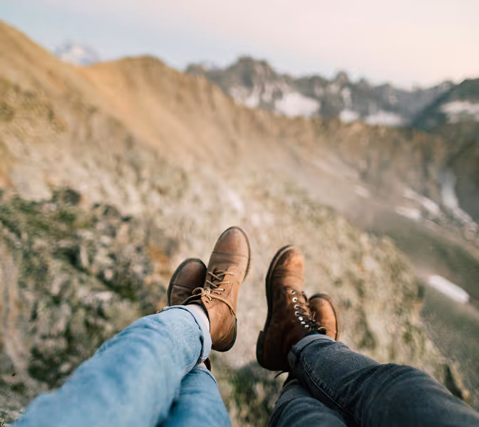 Two pairs of legs wearing boots resting with a mountainous landscape in the background.