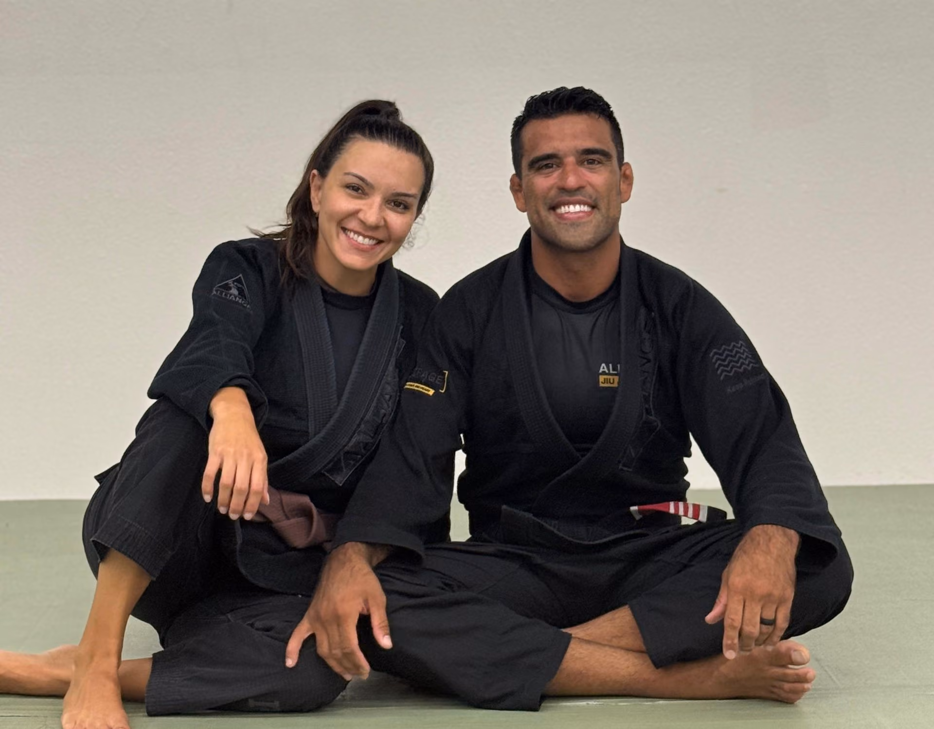 Smiling man and woman sitting cross-legged on mat wearing black Brazilian Jiu-Jitsu gis with belts.