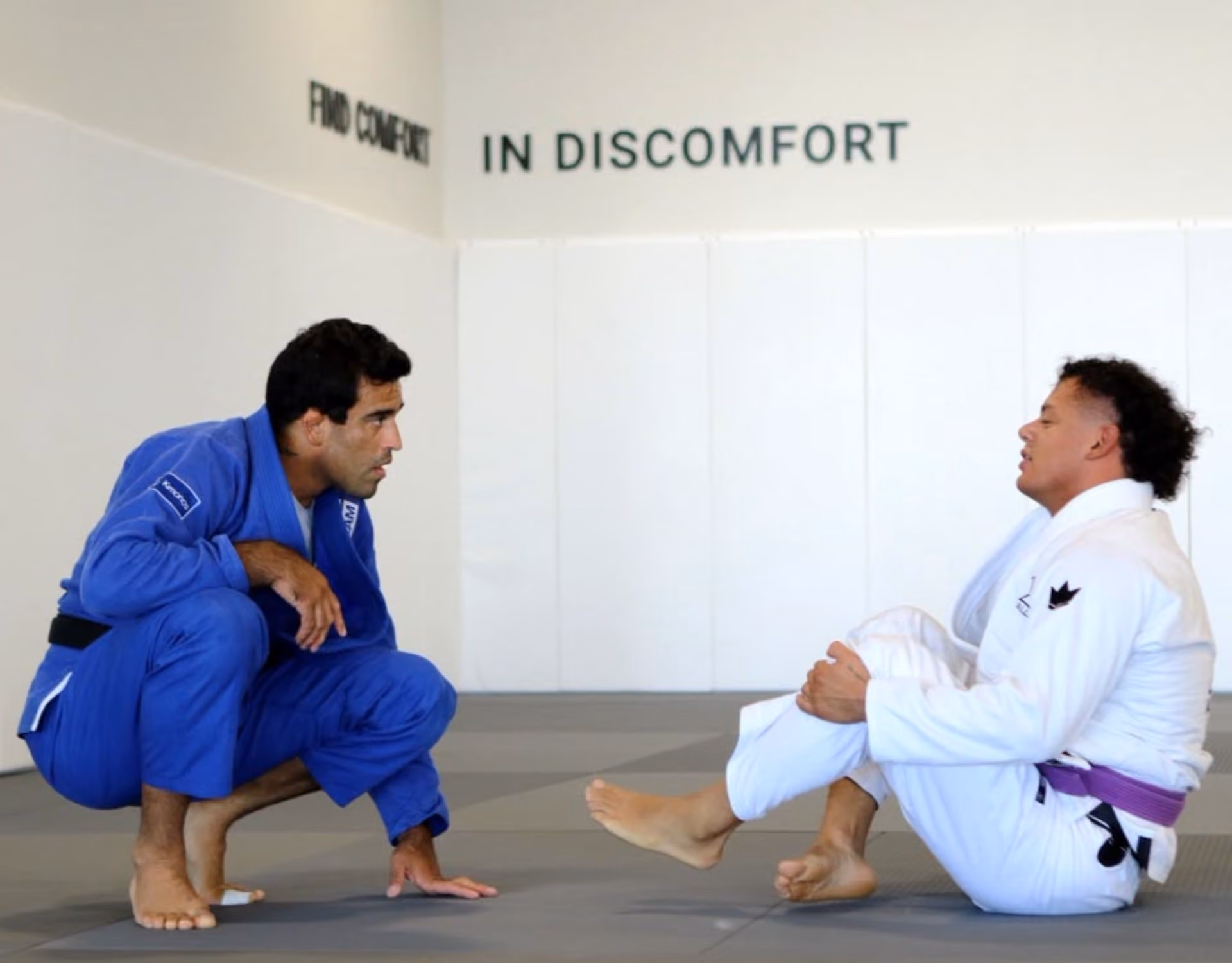 Two men in martial arts gis on mats; one crouches in blue, the other sits in white with a purple belt.