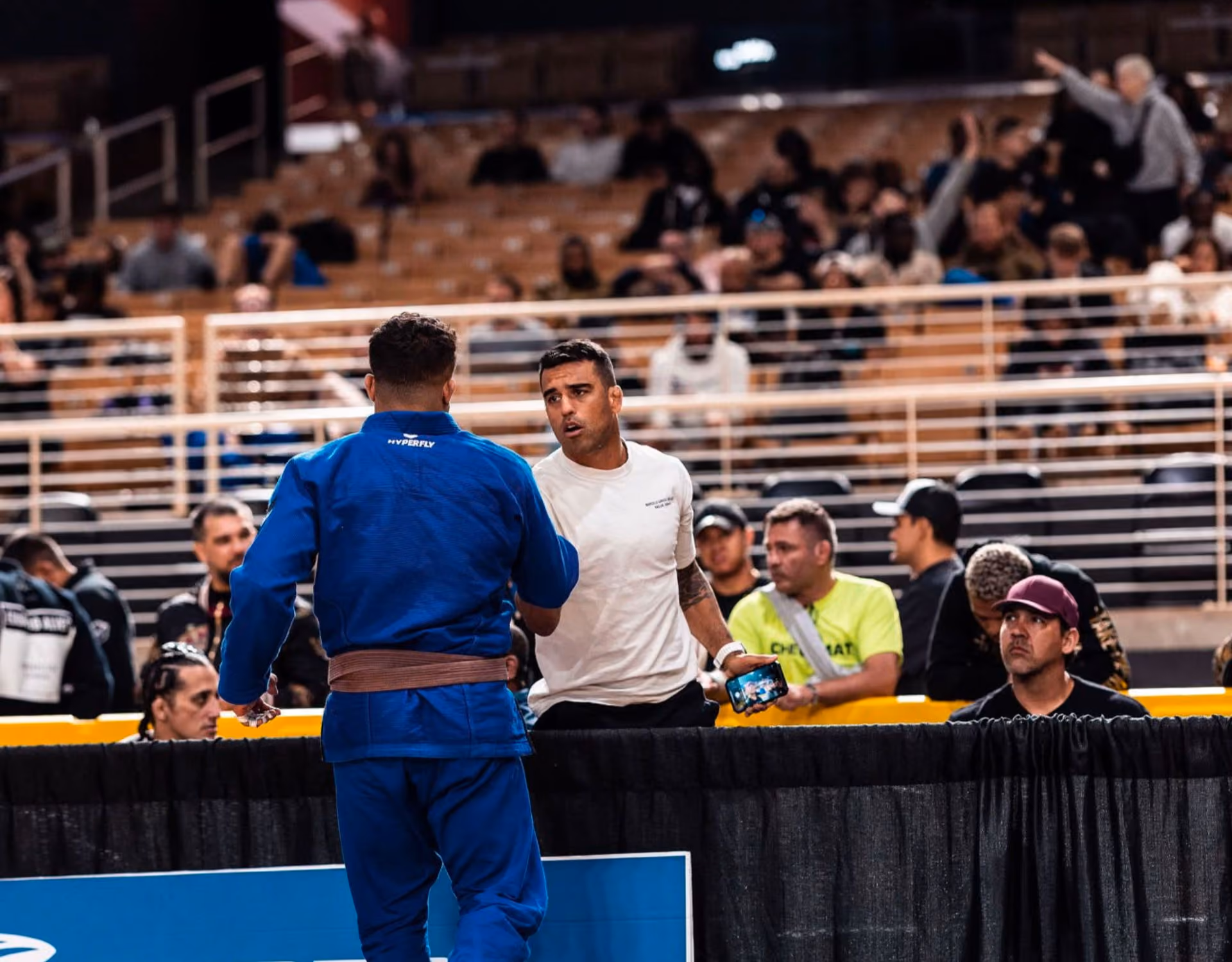 Man in blue martial arts gi shakes hands with man in white shirt holding a phone at a crowded event.