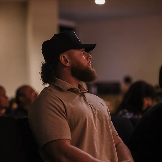 Bearded man in brown shirt and black cap seated in a dimly lit room, looking forward.