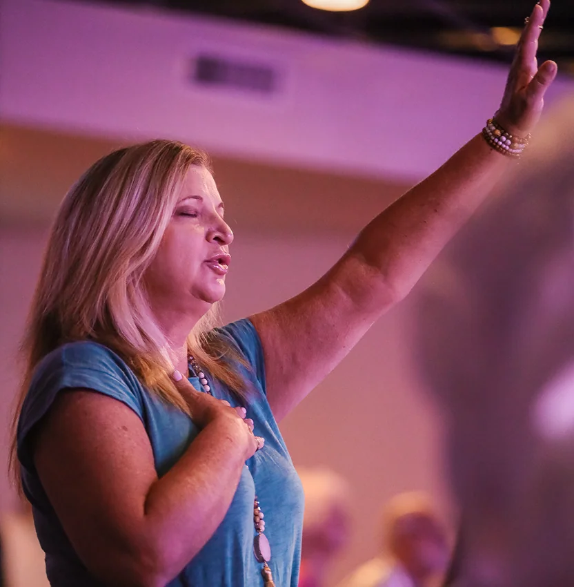 Woman with eyes closed singing and raising one hand during worship service.