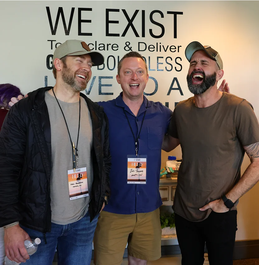 Three men laughing and posing with arms around each other in front of a motivational wall sign.