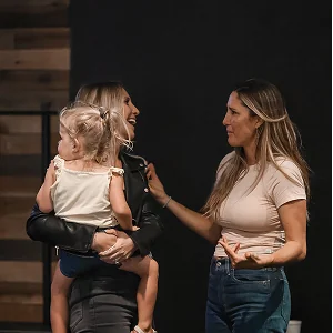 Two women smiling and talking, one holding a young child, in a casual indoor setting.