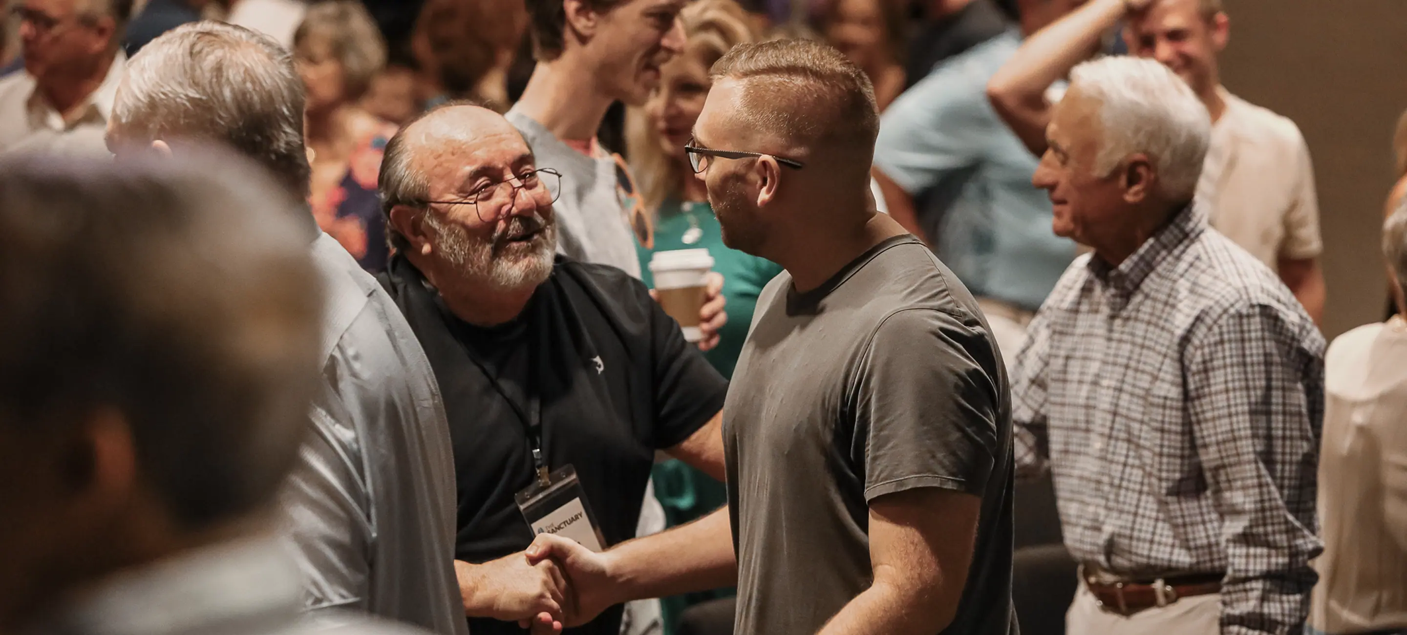 Two men smiling and shaking hands in a crowded social or conference setting.