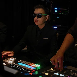 Man wearing dark sunglasses and suit operating a video switcher in a dim control room.