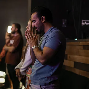 Man in blue shirt with hands clasped praying in a dimly lit room with others in background.
