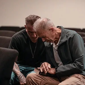 Two men seated close together, holding hands and praying with heads bowed in a quiet room.
