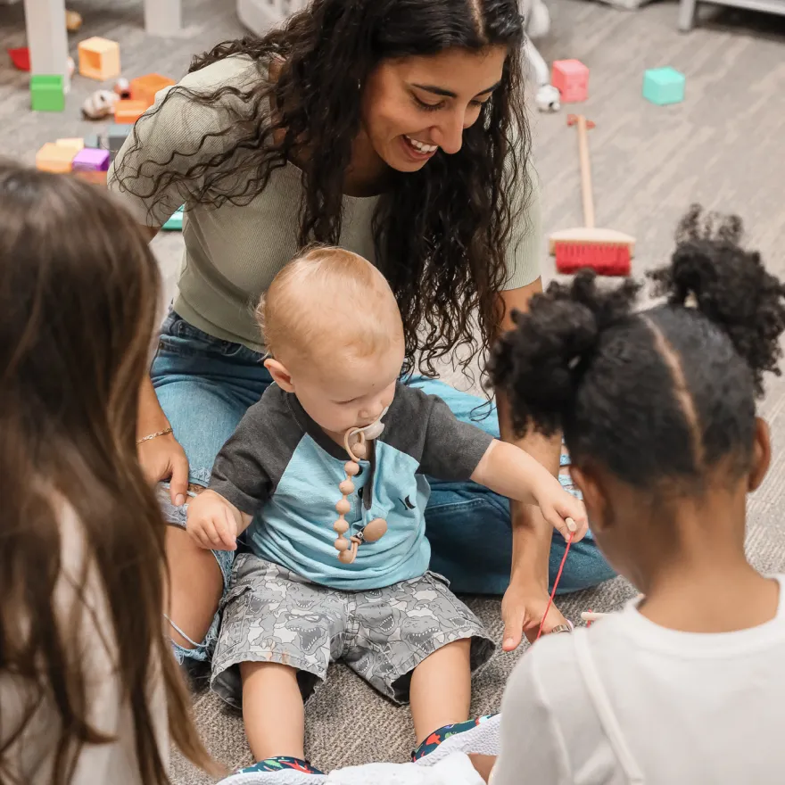 Woman sitting on floor playing with a baby and two children amid scattered toys indoors.