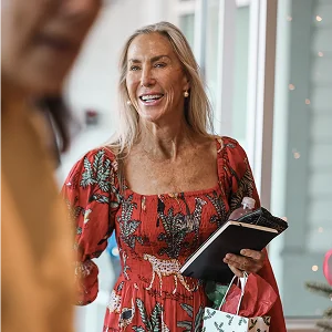 Smiling woman in red floral dress holding gifts and a notebook indoors.