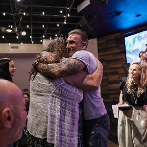 Smiling man with tattoos hugging woman in a warmly lit indoor gathering with other people nearby.