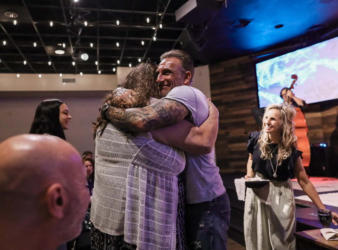 Man and woman hugging warmly at an indoor event with others smiling nearby.