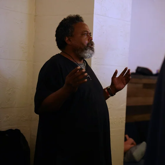 Man with gray beard speaking and gesturing with his hands against a beige wall.