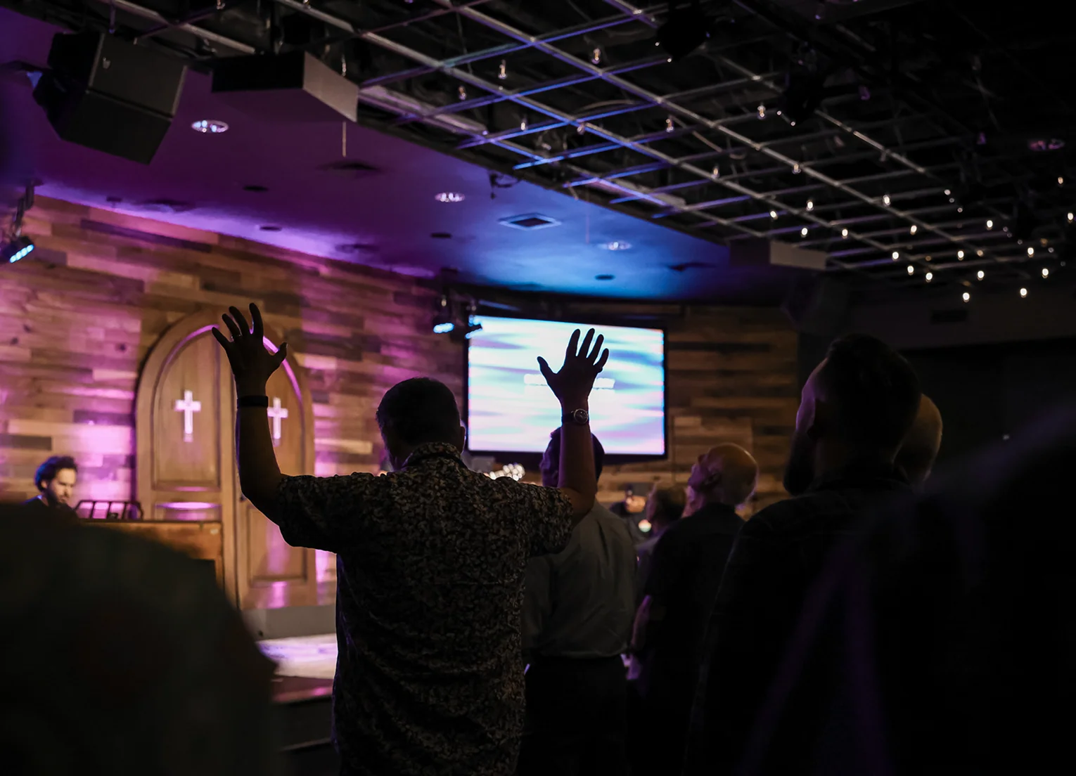 Congregation worshiping indoors with raised hands and crosses lit on wooden walls.