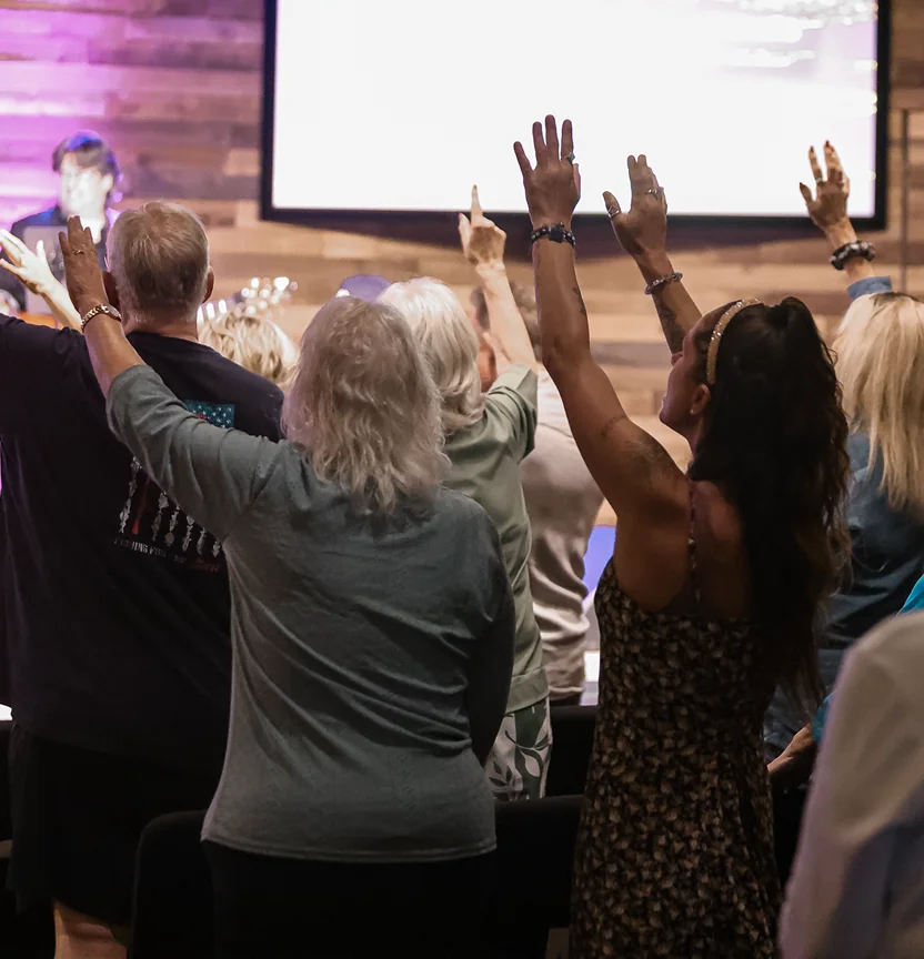Diverse people raising hands during a worship service in a wood-paneled room with a large screen.