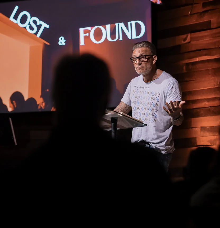 Man with glasses speaking at a podium with a screen behind showing words LOST & FOUND.
