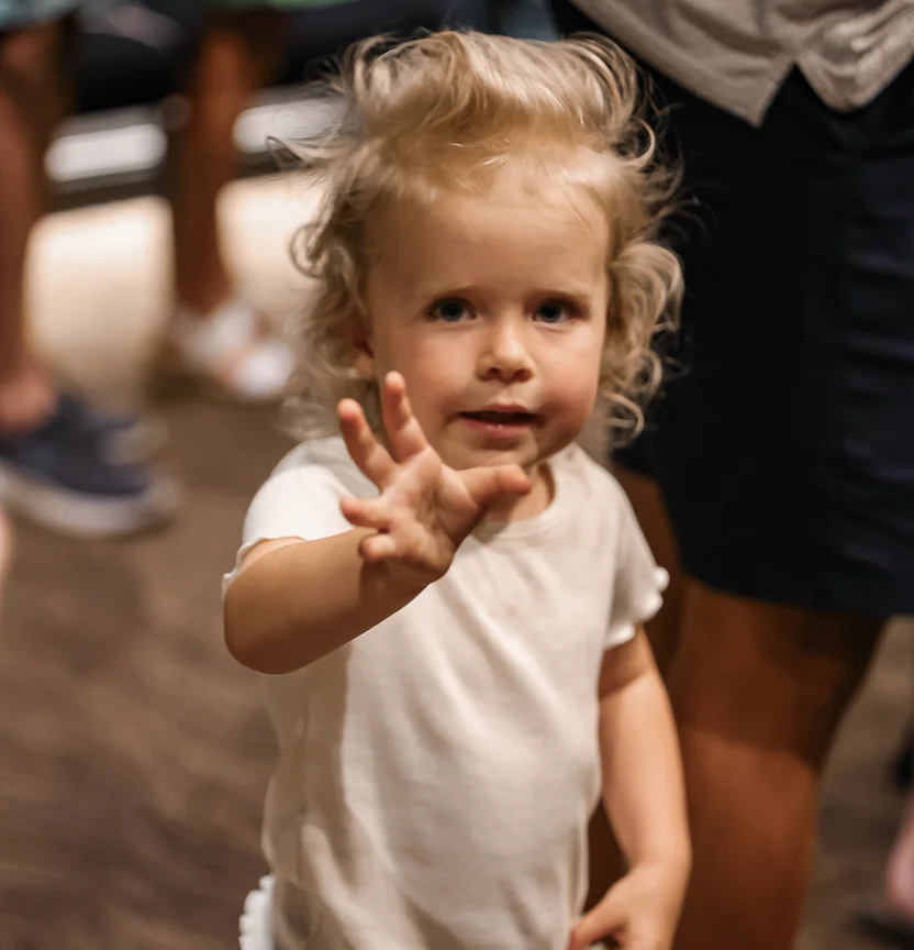 Young child with curly blonde hair wearing a white shirt, waving with one hand.