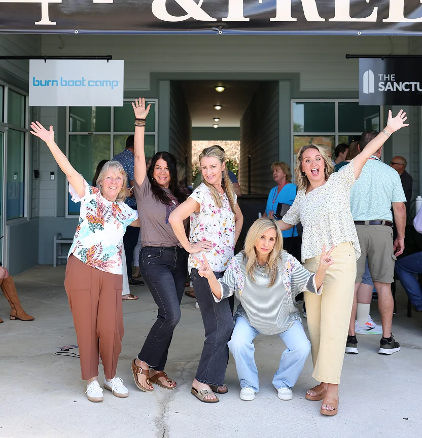 Five women smiling happily with arms raised outside a building with Burn Boot Camp signs.