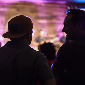 Two men seen from behind at a nightclub event with purple lighting and a blurred performer on stage.