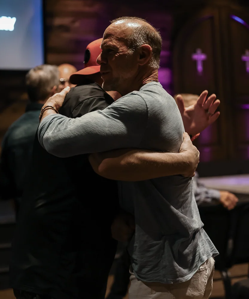 Two men warmly hugging in a dim church setting with crosses in the background.
