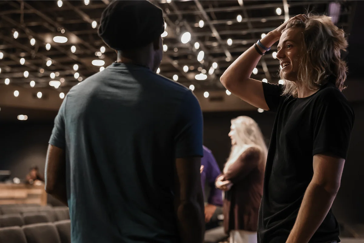 Two people talking in a dimly lit room with hanging lights and rows of seats in the background.
