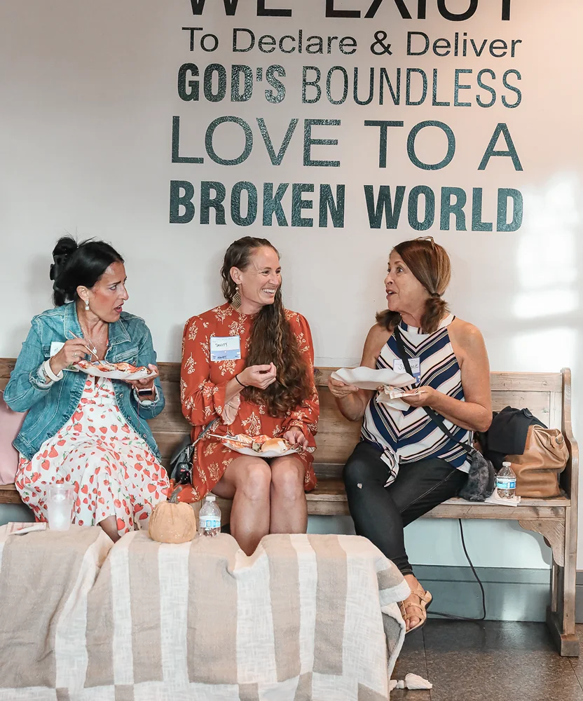Three women sitting on a bench eating and chatting beneath a wall sign about God's boundless love.