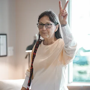 Middle-aged woman with glasses making a victory sign indoors by a window.