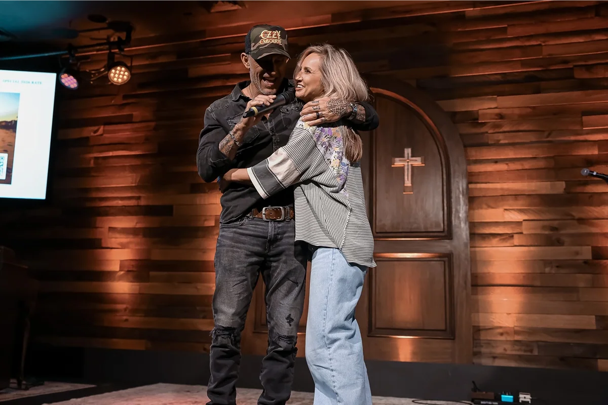 Man and woman hugging on a stage with wooden background and a cross on a door behind them.