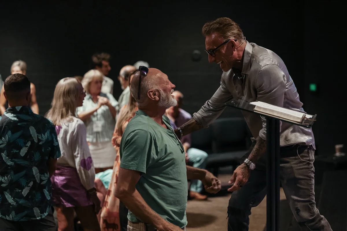 Man in glasses speaking and smiling warmly to a seated bald man in green shirt at a gathering.