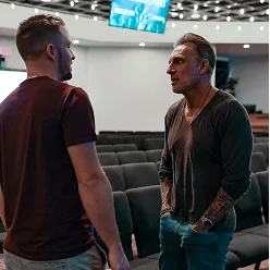 Two men standing and talking in a modern auditorium with rows of seats and a ceiling screen.