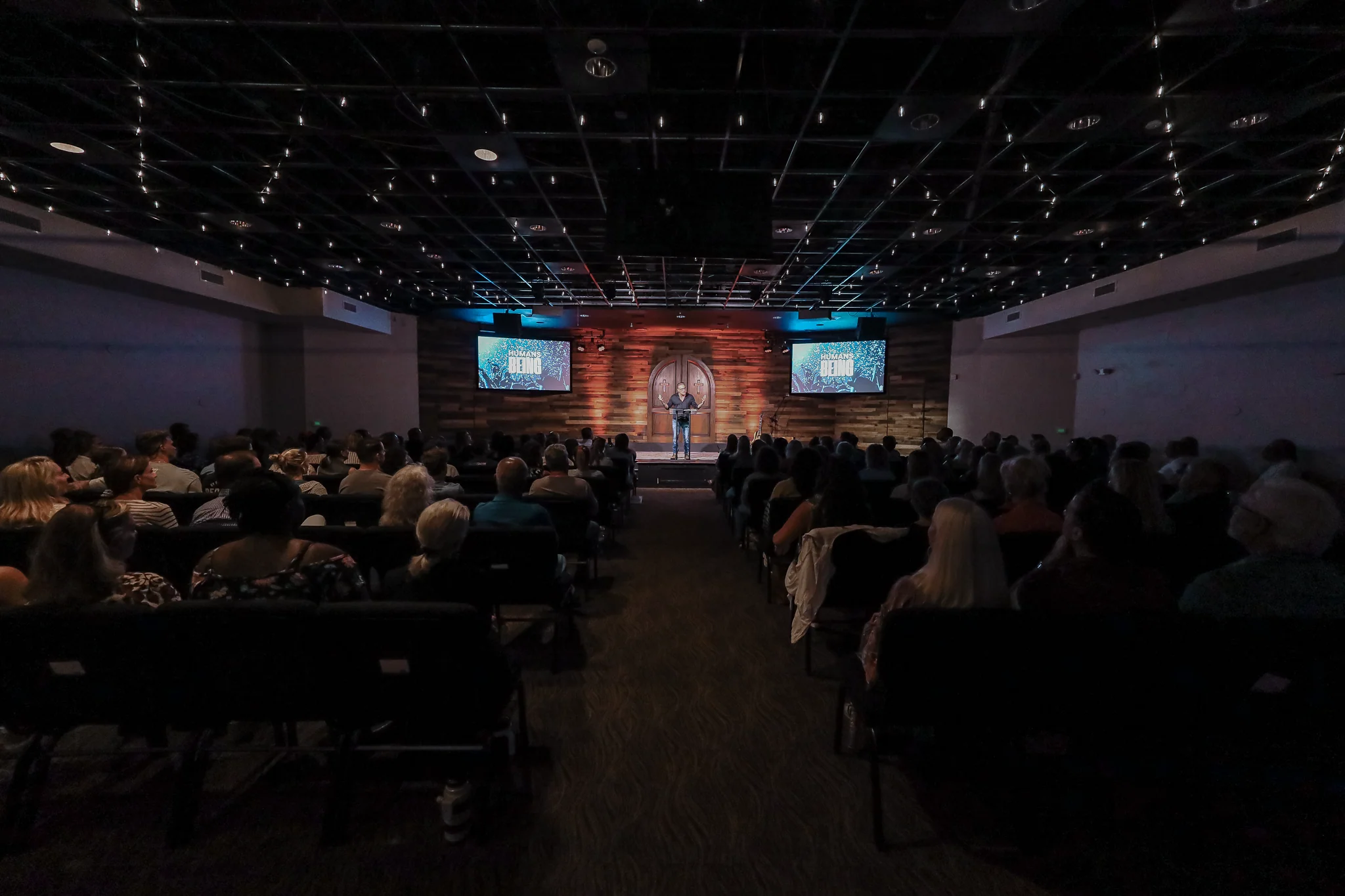 A speaker stands at a podium in a dim auditorium filled with seated audience members.