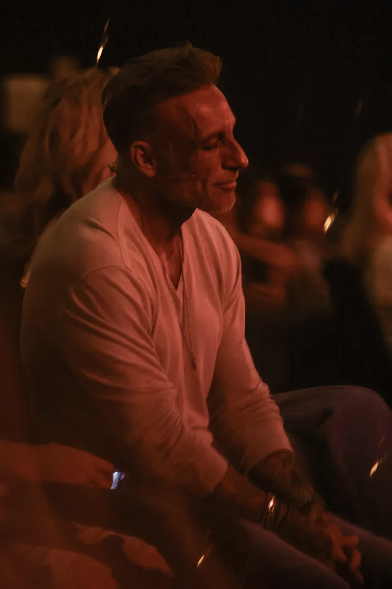 Man in a white long-sleeve shirt sitting with eyes closed and hands clasped in a dimly lit setting.