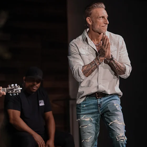 Tattooed man in a light shirt and ripped jeans praying on stage with a seated man playing guitar behind him.