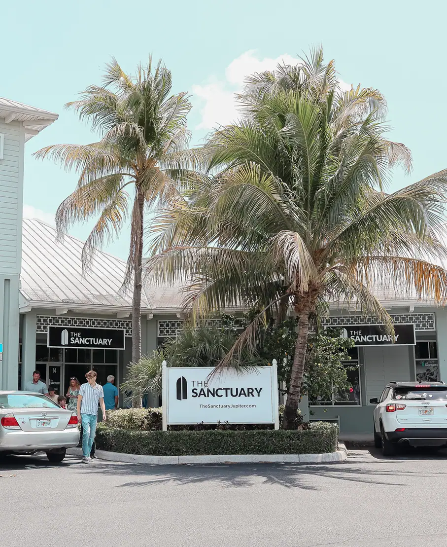 Entrance to The Sanctuary with palm trees, people, and parked cars in a sunny parking lot.