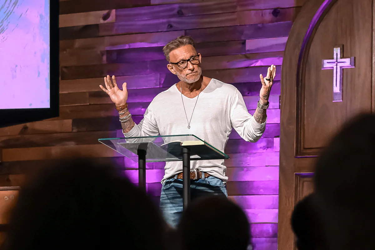 Man wearing glasses speaks at a church podium with a cross-lit wooden wall behind him.