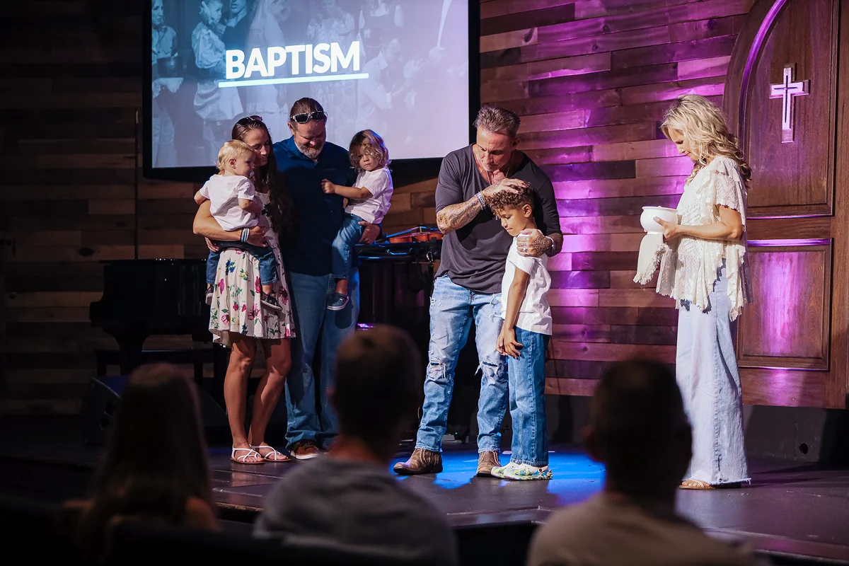 Group on stage during a baptism event, with a man blessing a child and family standing nearby.