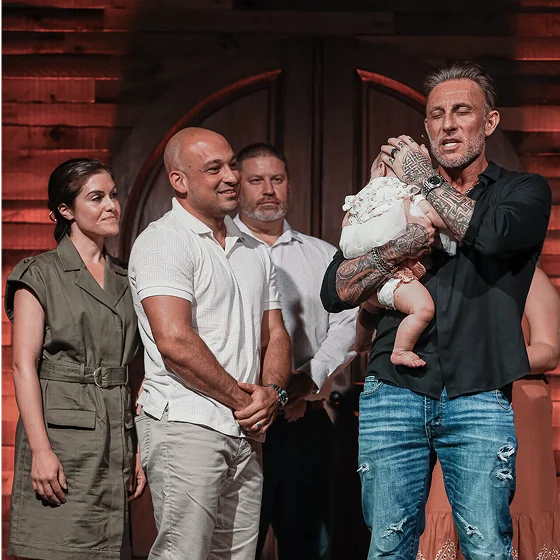Man holding baby with eyes closed, three adults standing nearby in a warmly lit indoor setting.