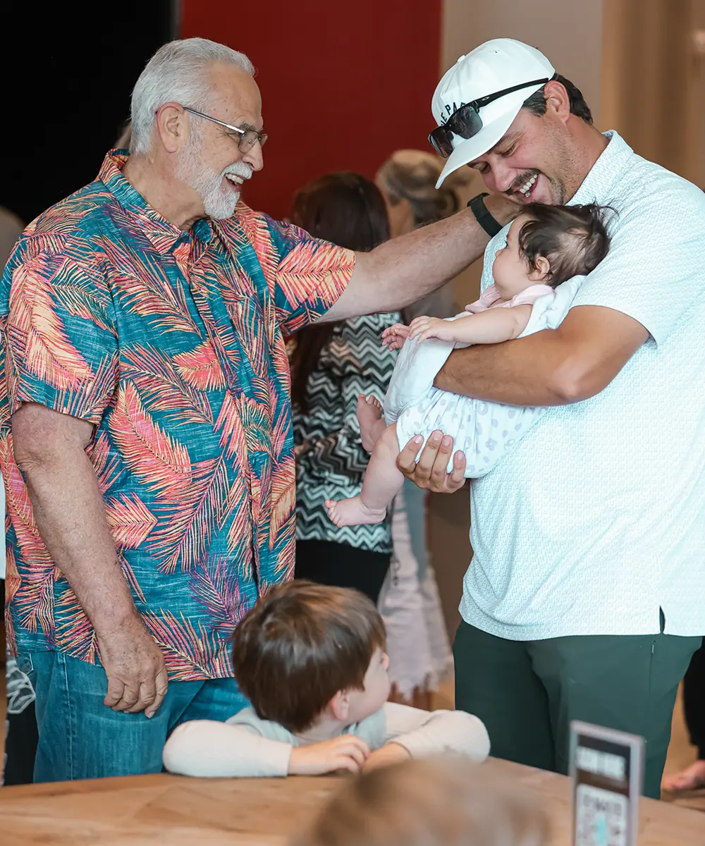 Smiling elderly man touches shoulder of man holding a baby while a child watches at a table.