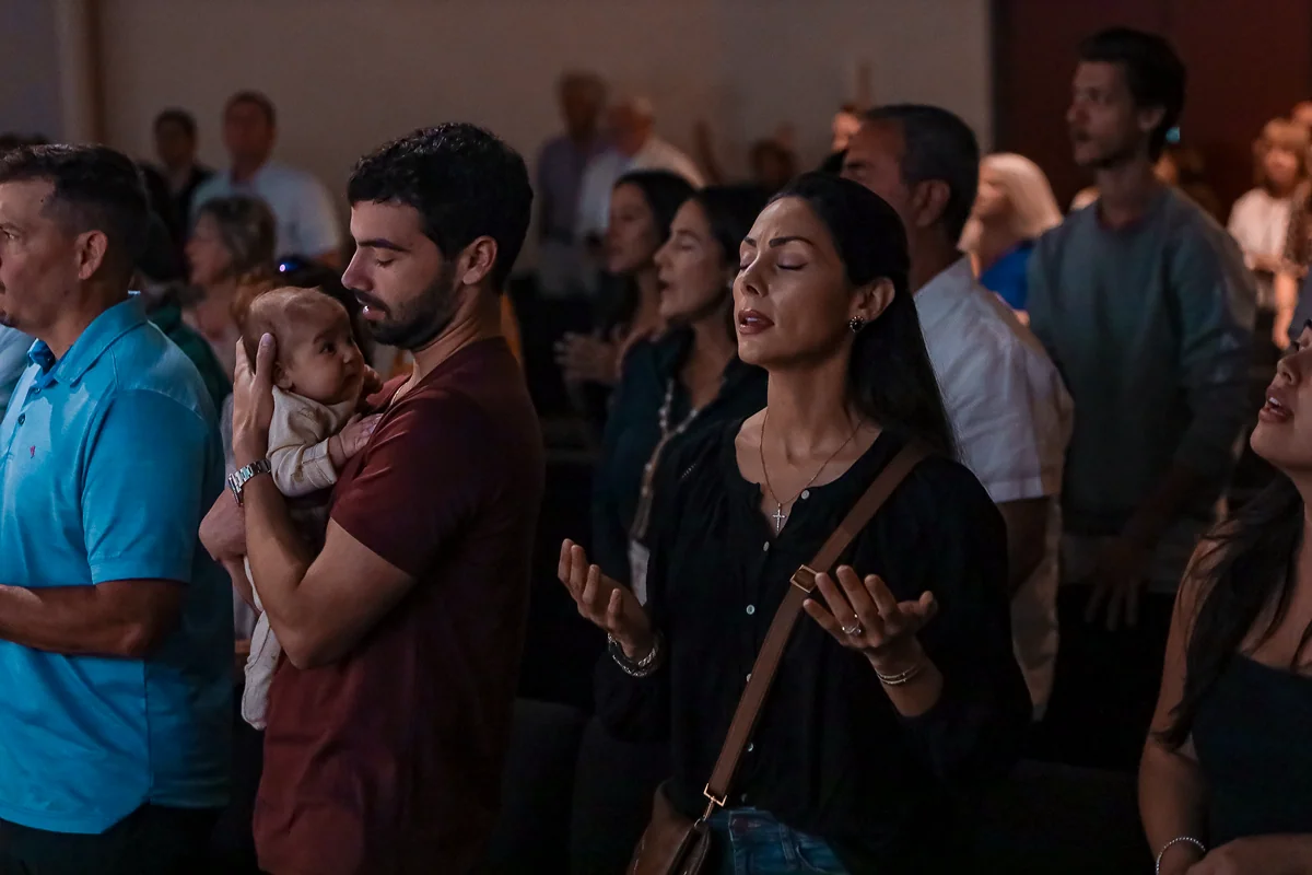 People standing with eyes closed and raised hands in a moment of prayer or worship indoors.