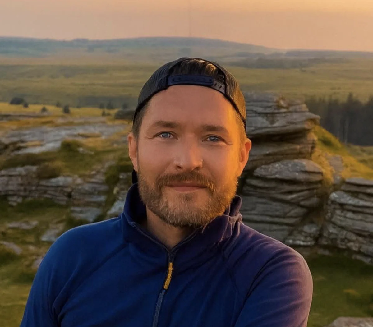 Man with beard wearing a blue jacket and black cap outdoors with rocky landscape in background.