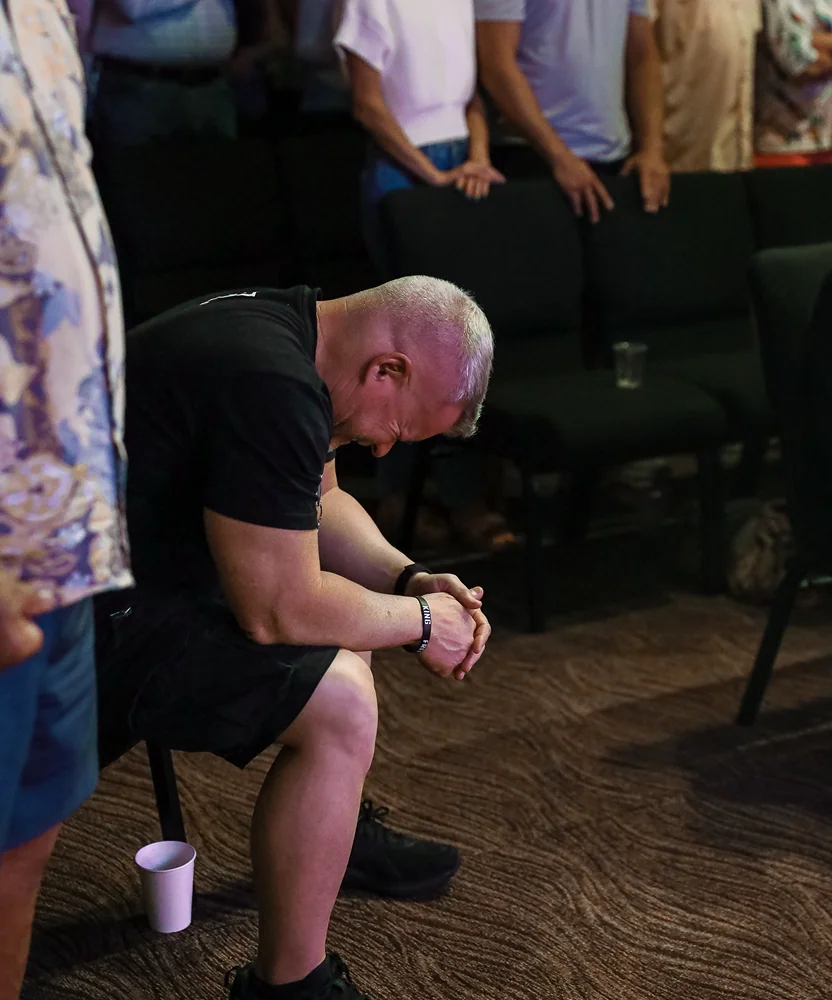 Man in black clothes sitting and praying with head bowed in a room with seated people.