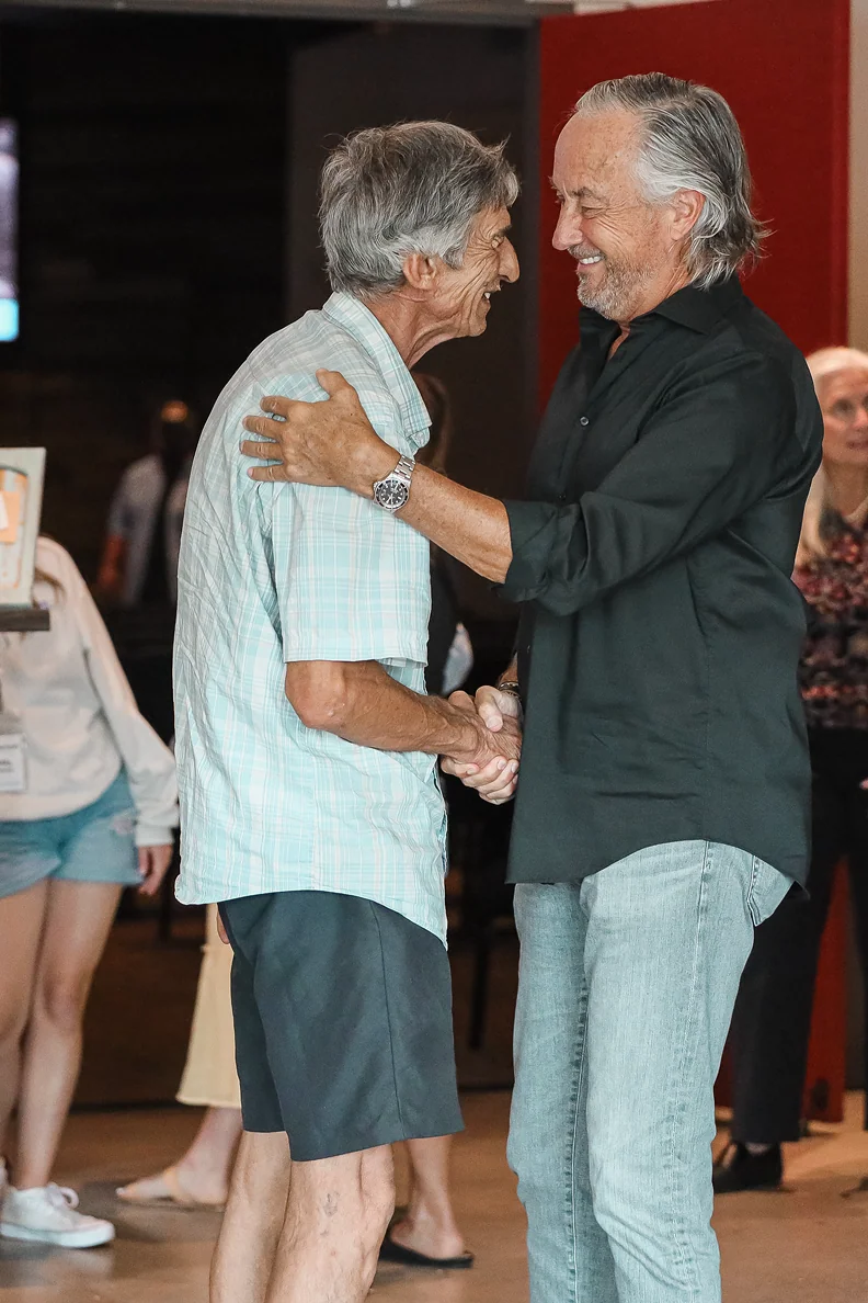 Two older men smiling warmly while shaking hands and embracing indoors.