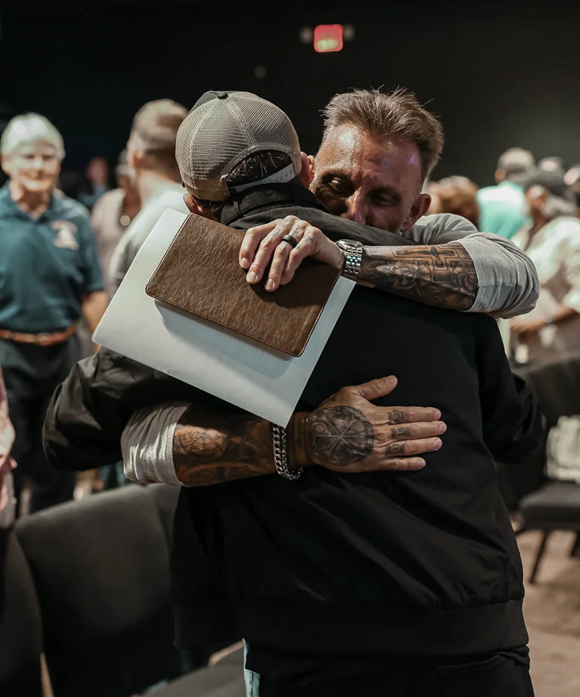 Two men warmly embracing, one holding a Bible and a card, in a gathering with people in the background.
