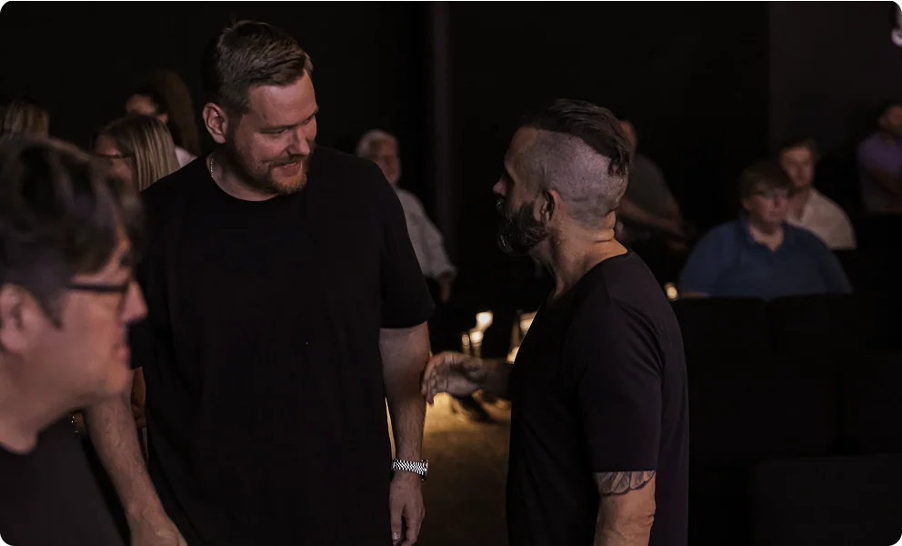 Two men in black shirts conversing in a dimly lit room with seated audience in the background.