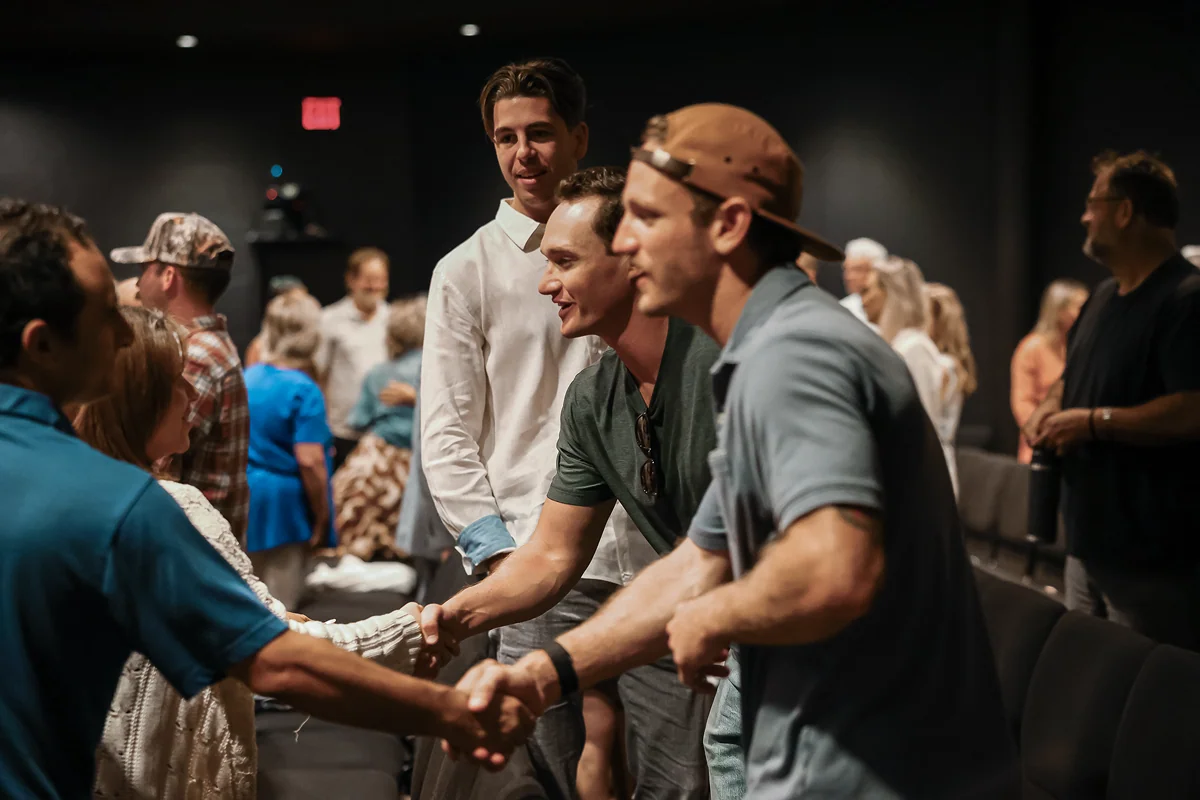 People shaking hands and greeting each other indoors, with others socializing in the background.