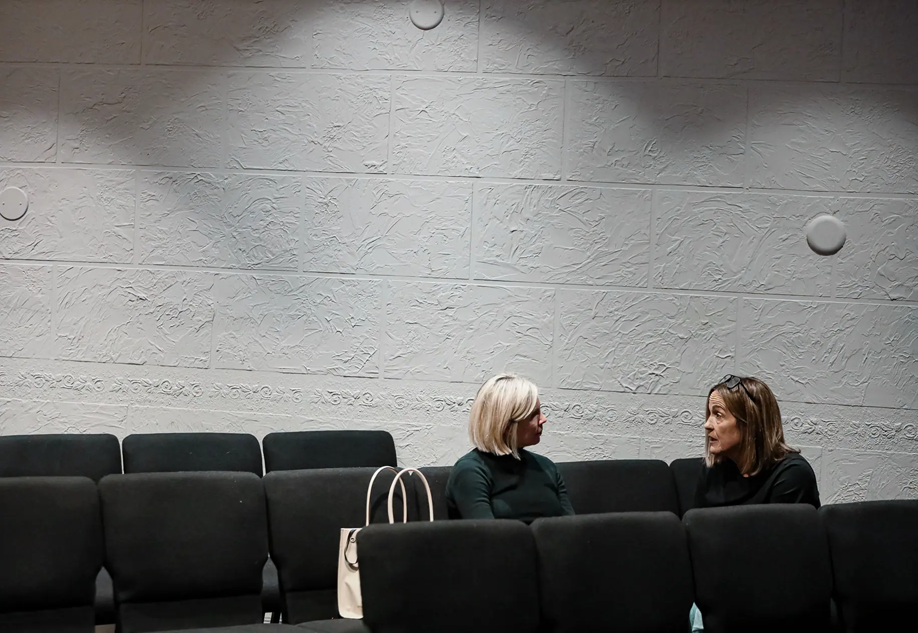 Two women sitting and talking in a mostly empty room with textured gray walls and black chairs.