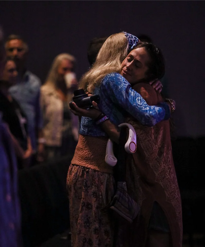 Two women warmly hugging in a dimly lit room with blurred people in the background.