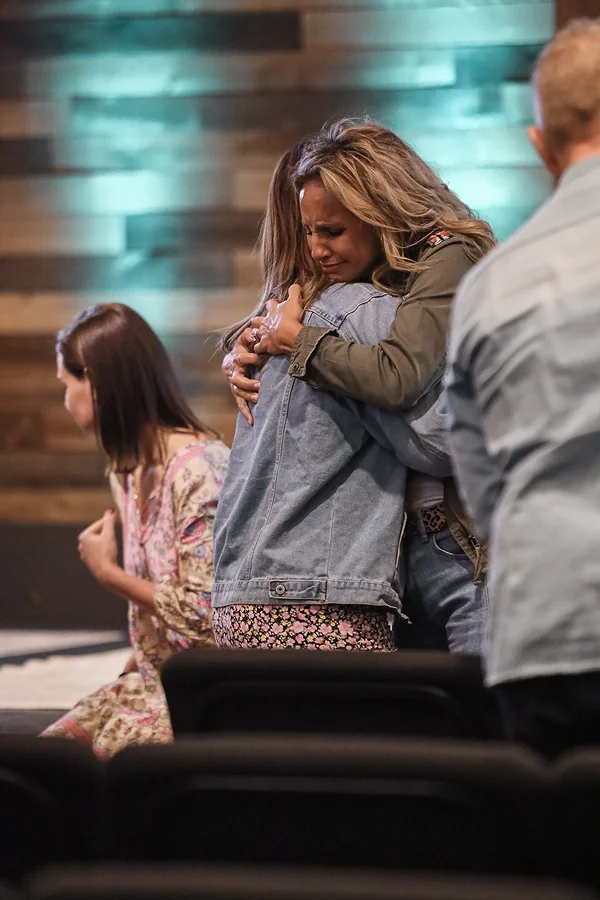 Two women embrace tightly, one visibly emotional, inside a church service setting with wooden wall background.